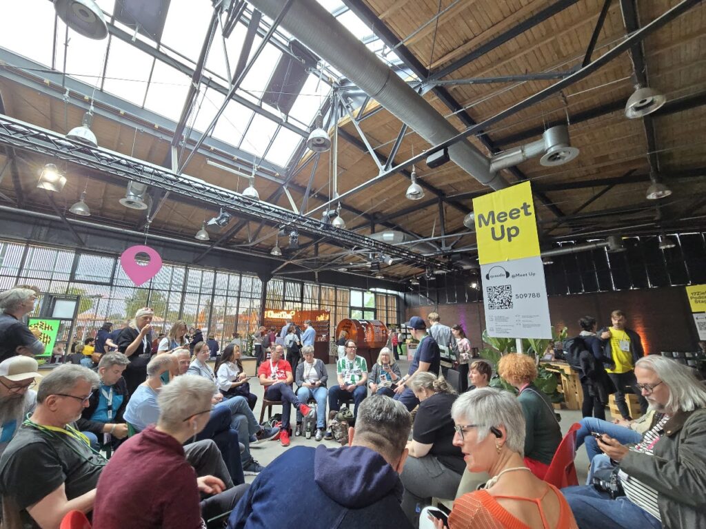 ine große Gruppe von Menschen sitzt und unterhält sich in einer großzügigen Halle im Industriestil während eines Community-Meetups.