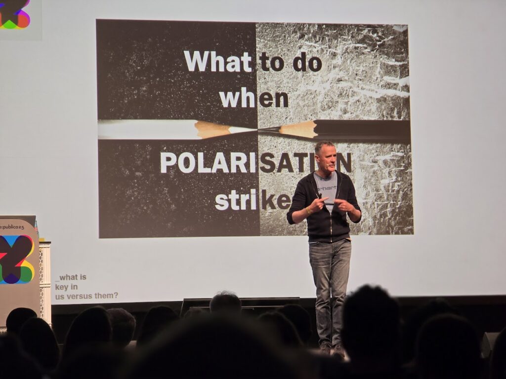 Ein Mann spricht auf einer Bühne vor einer Leinwand mit der Folie „What to do when POLARISATION strikes“ und dem Bild zweier zerbrochener Bleistifte.