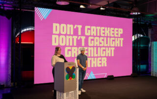 Zwei Personen stehen auf einer Bühne vor einer großen, pinken Leinwand mit der Botschaft: „DON’T GATEKEEP, DON’T GASLIGHT, GREENLIGHT GATHER.